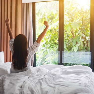 Woman waking up in bed with sun shining through windows.